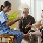 An elderly couple happily receives care information at home from a trained female nurse for a family member who is sitting in a wheelchair.