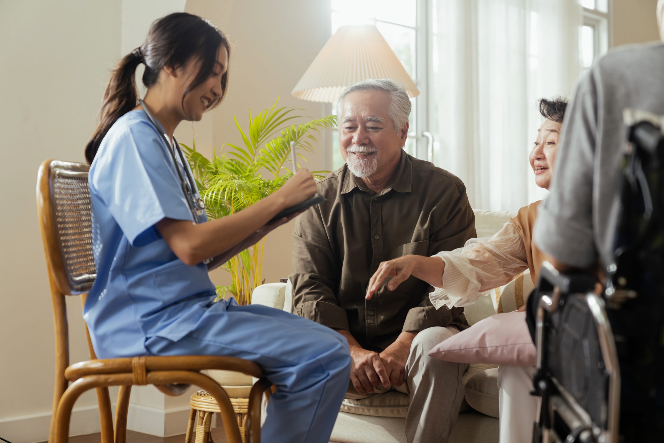An elderly couple happily receives care information at home from a trained female nurse for a family member who is sitting in a wheelchair.
