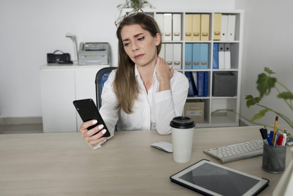 An European young women sitting inside her corporate office room looking at her mobile as part of her everyday work and holding her paining shoulder. Pain caused by poster is visible on her face.